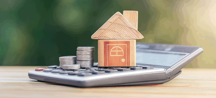 A wooden house model placed on a calculator with stacks of coins beside it, representing financial planning and investment in real estate.
